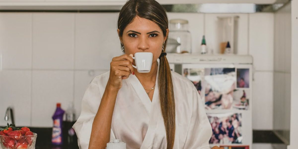 A woman in a white robe enjoys coffee with breakfast in a cozy kitchen setting.